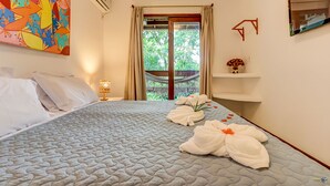 A hotel room with a bed, a window with curtains, a wall-mounted air conditioner, a decorative painting, and a small shelf with a vase. -  Pousada Vila Galea (Itacaré)