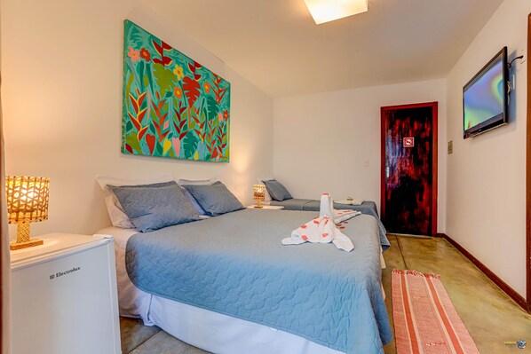 A bedroom with a bed, a nightstand, a lamp, a painting, a television, and a door. - Pousada Vila Galea (Itacaré)