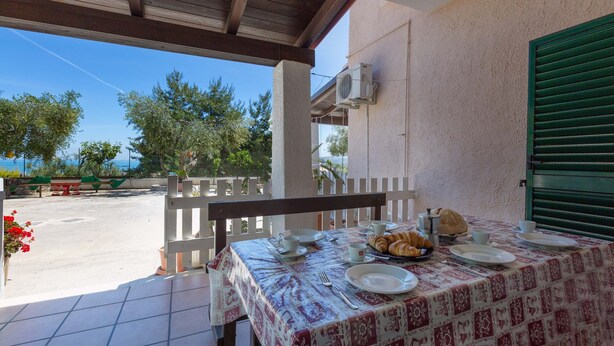 A covered patio with a table set for breakfast, outdoor seating, and a view of trees and a fence.