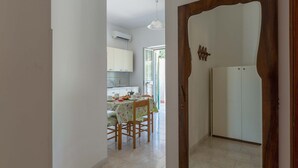 A narrow hallway leading to a kitchen with a dining table and chairs, a white refrigerator, and a wooden door frame.