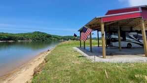 A lakeside RV park with a covered pavilion, an American flag, and a white RV parked under the pavilion. - Reconnect with nature at this unforgettable escape (RV NOT INCL'D). (Clifton)