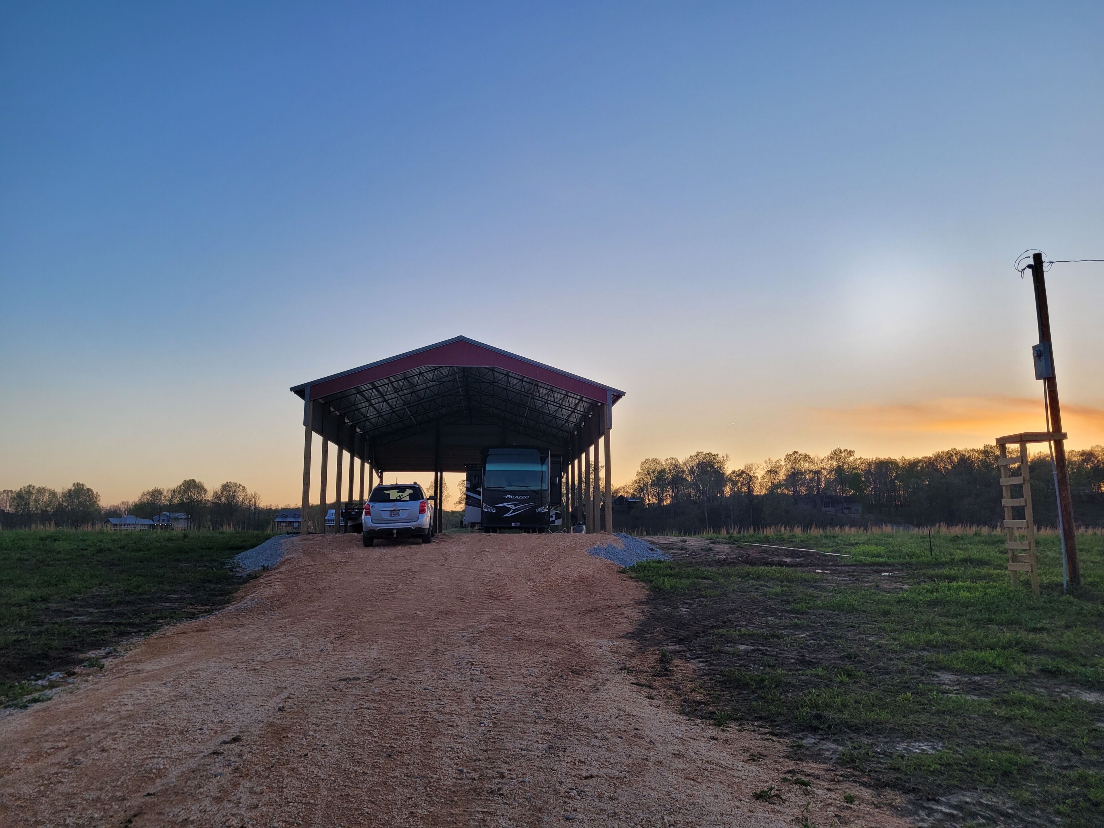 A covered outdoor area with a dirt path leading to a bus and a car parked under a metal roof structure.