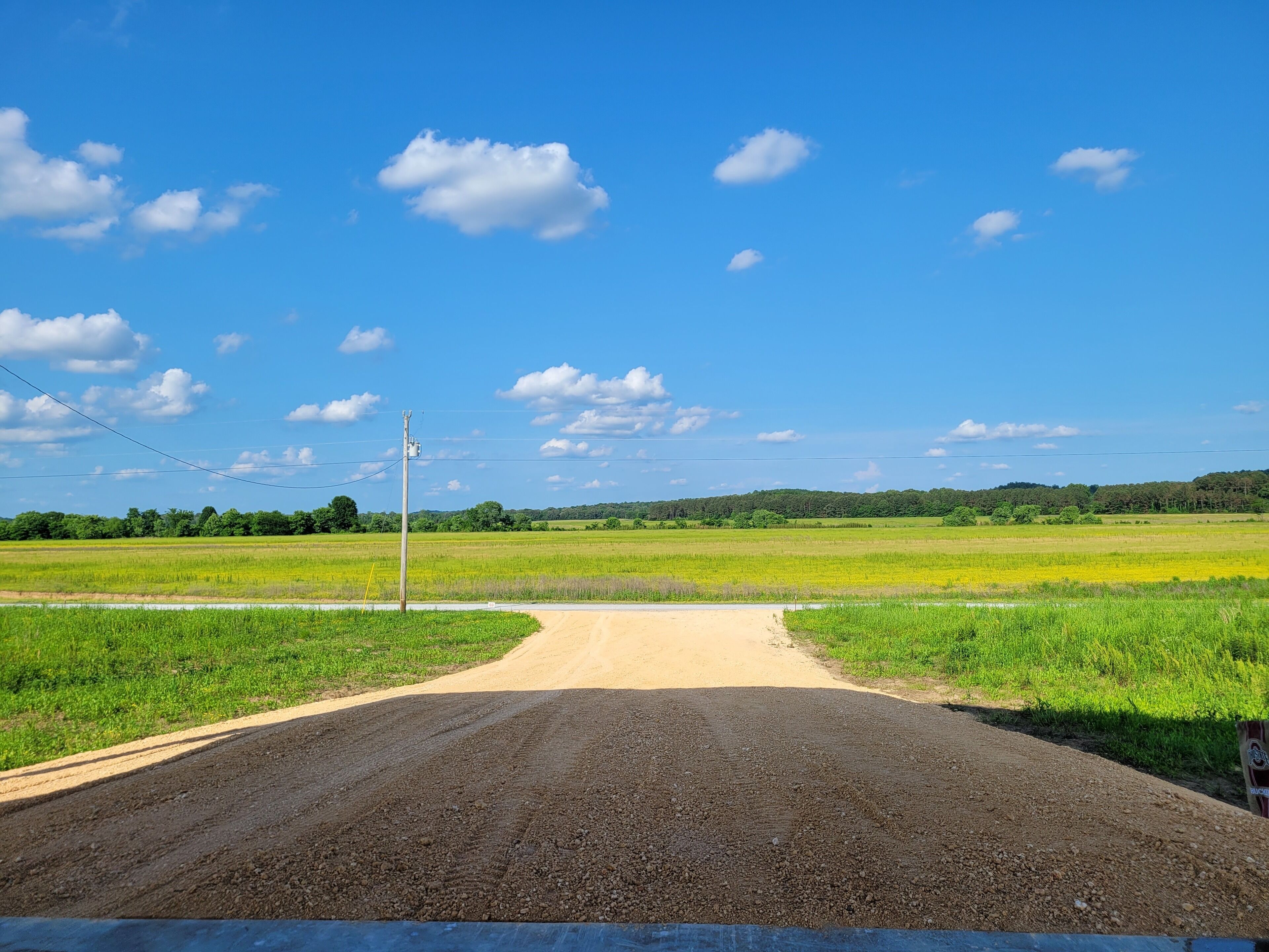 A rural road leading to a green field with a utility pole and power lines.