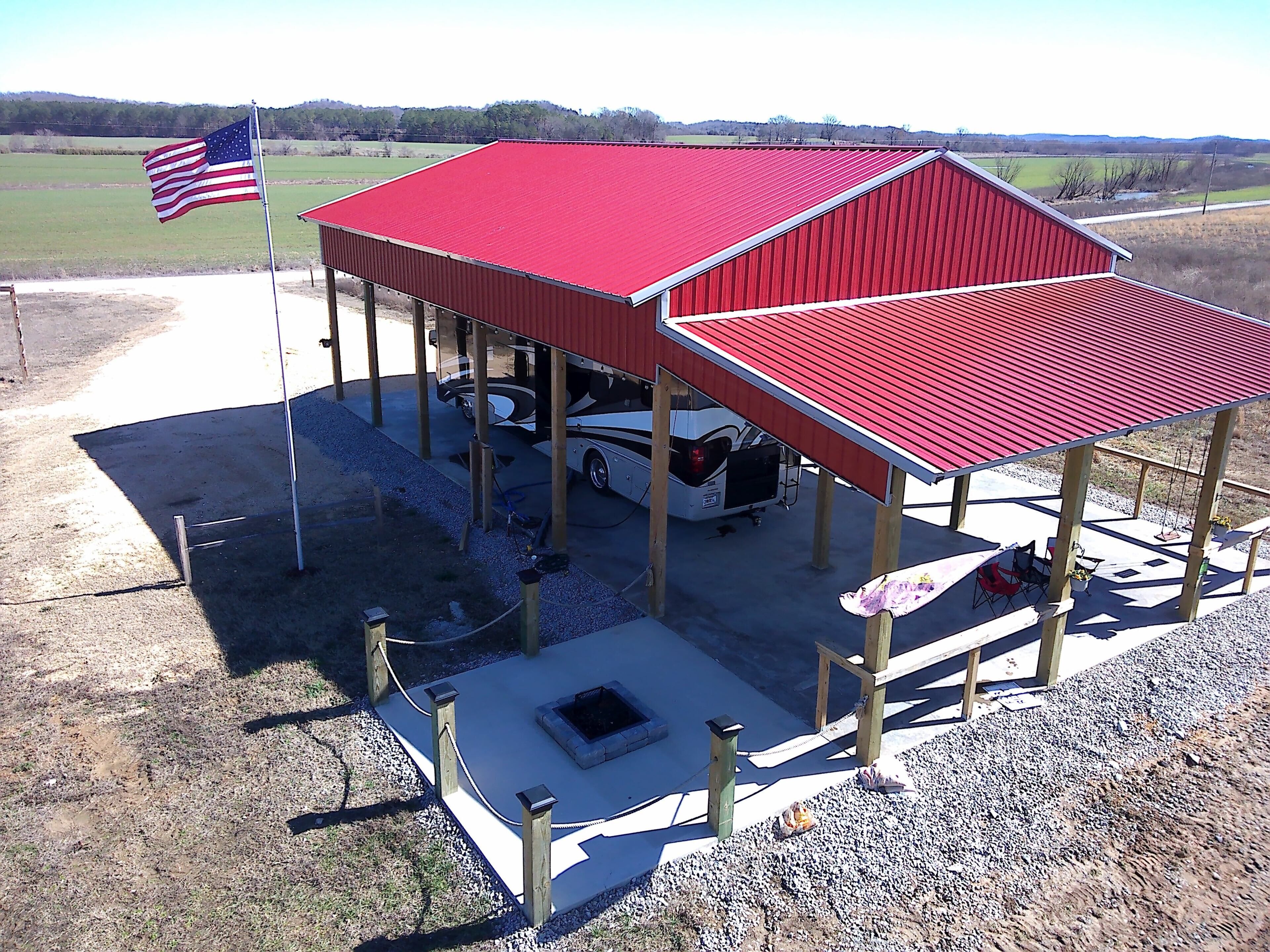 A covered outdoor area with a red roof, a flagpole with an American flag, and a white RV parked underneath.