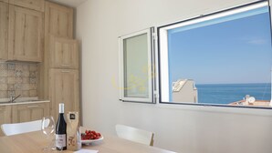 Une cuisine avec des meubles en bois, une table à manger avec une bouteille de vin, un saladier de fruits et une fenêtre donnant sur la mer.