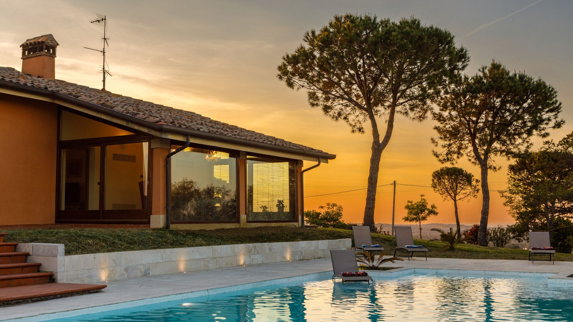 A modern villa with a swimming pool, large windows, and a view of the sunset.