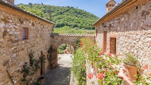 A stone building with a narrow passageway, a rustic archway, and a view of a hill with greenery and a tower in the distance. - Podere Pratovecchio 14+2, Emma Villas (Montefollonico)