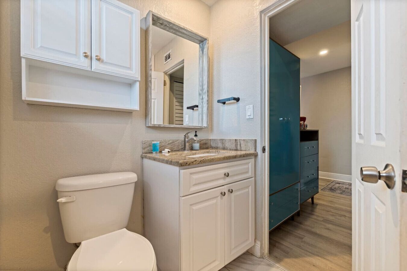 A bathroom with a toilet, a sink with a marble countertop, a mirror above the sink, and a teal cabinet.