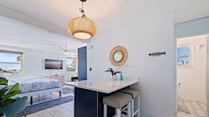 Une cuisine moderne avec un îlot central, un coin repas agrémenté d’une chaise bleue, et une chambre avec vue sur la mer.