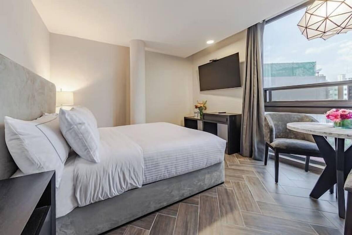 Ein modernes Hotelzimmer mit einem großen Bett, einem Schreibtisch mit Flachbildfernseher, einem Esstisch mit Stühlen und einem großen Fenster mit Blick auf die Stadt.