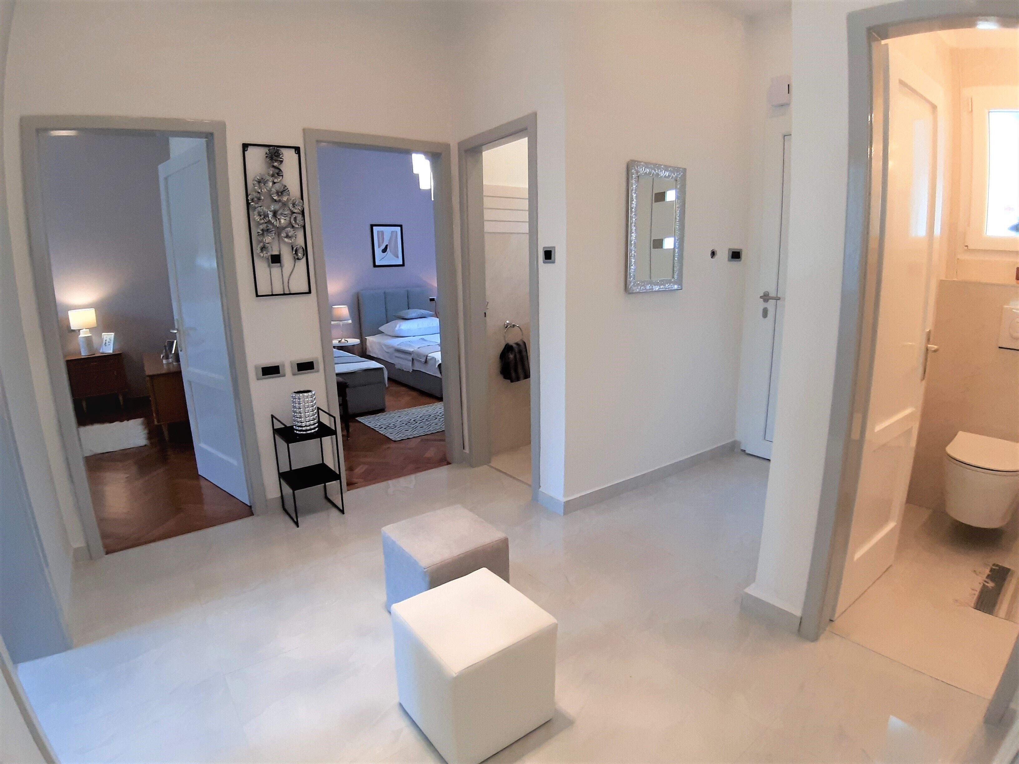 Une chambre d’hôtel moderne dotée d’un grand lit, d’un coin salon avec une petite table et un pouf, d’une salle de bain avec des toilettes et un lavabo, et d’un couloir menant à une autre chambre.