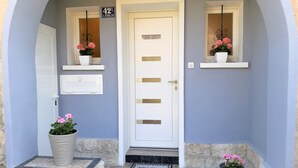 Entrada de una casa con una puerta blanca, un buzón y dos macetas con flores rosas.