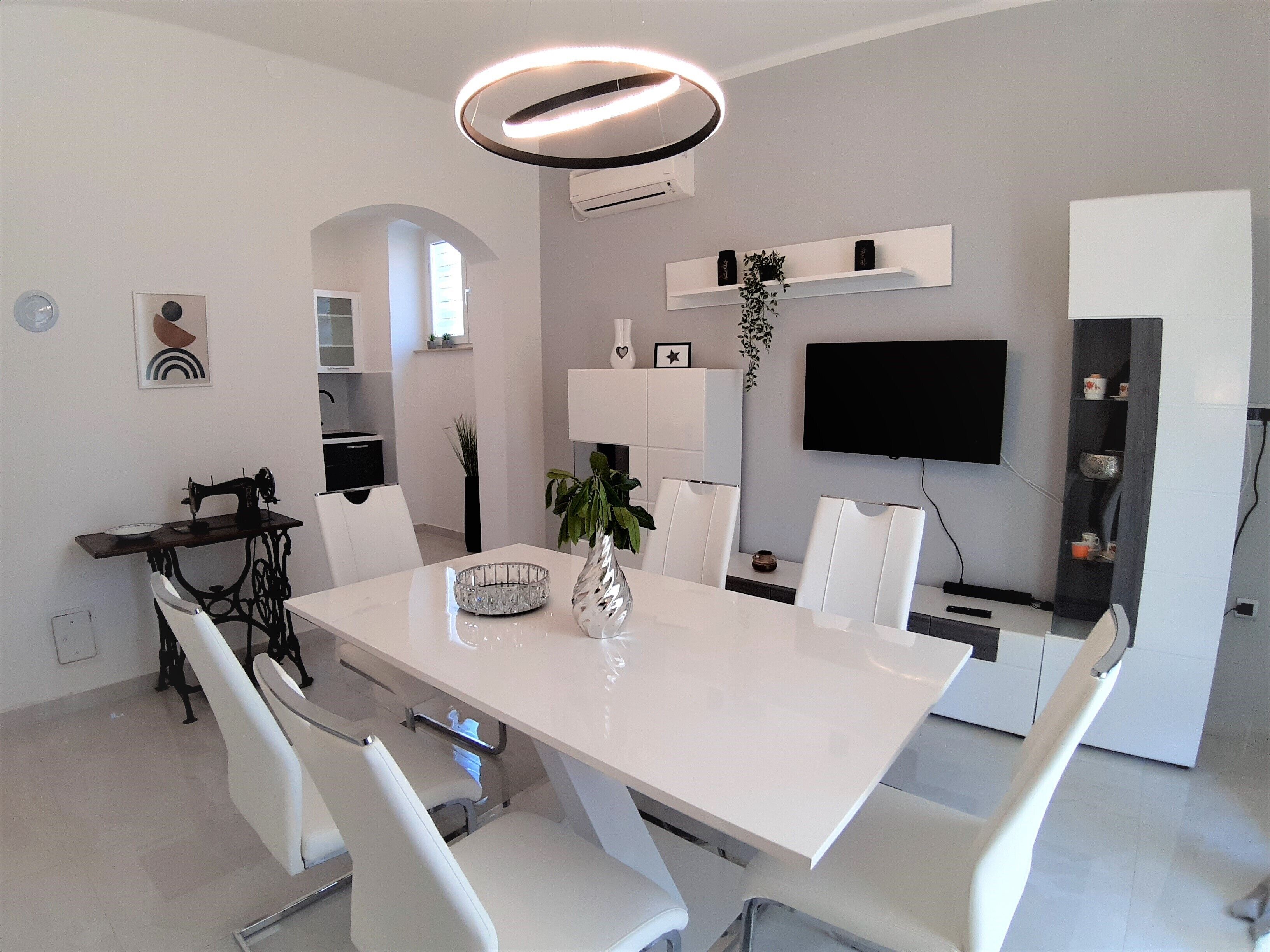 A modern dining area with a white table, chairs, and a chandelier. There is a television mounted on the wall and a plant on a shelf.