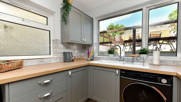 A compact kitchen with grey cabinets, a wooden countertop, a washing machine, and a window overlooking a patio area. - Just Stay Wales - Hartwell House Mumbles (Swansea)