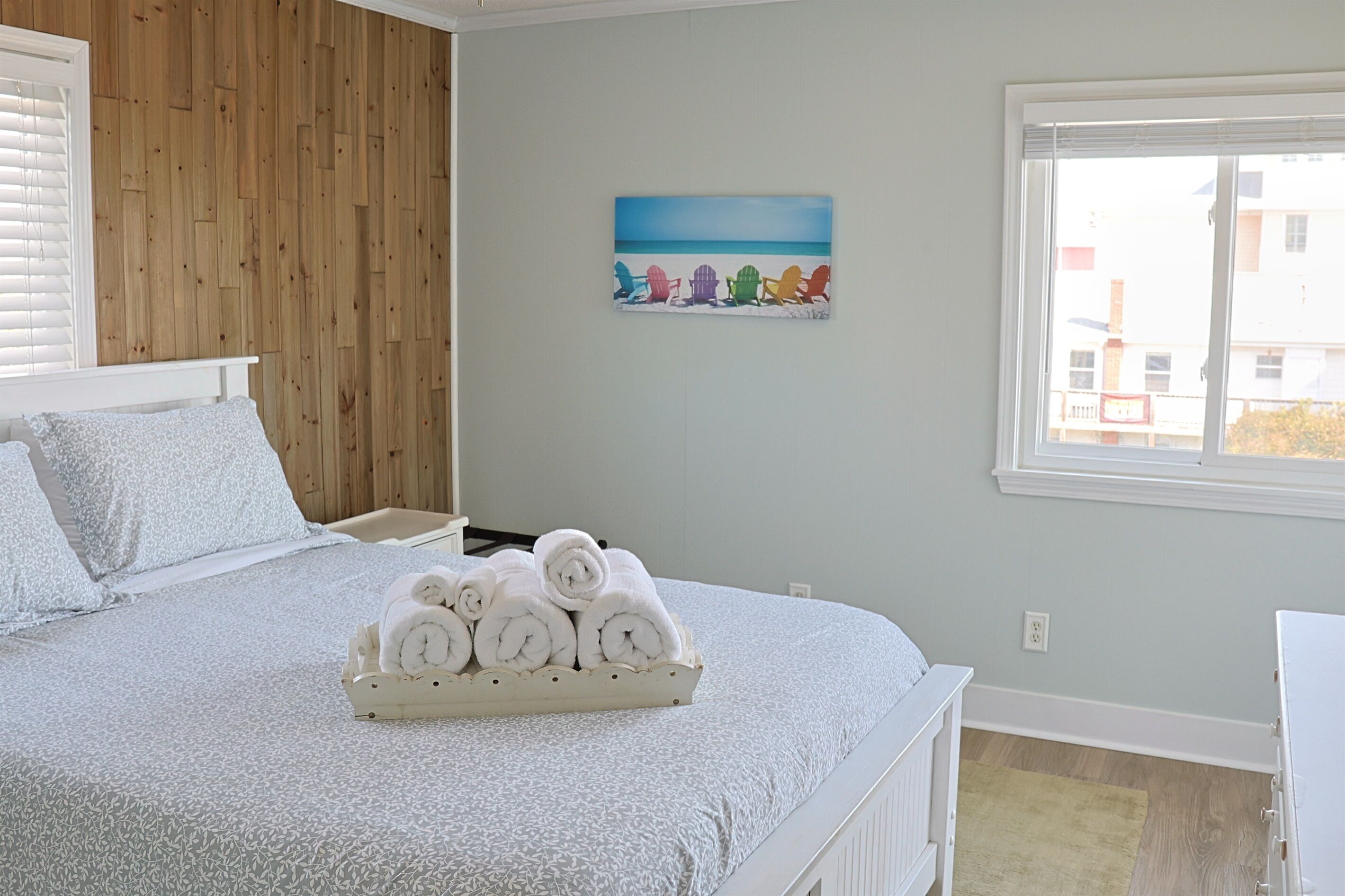 Une chambre à coucher avec un lit, un panneau mural en bois, un tableau représentant des chaises de plage et une fenêtre donnant sur un paysage.