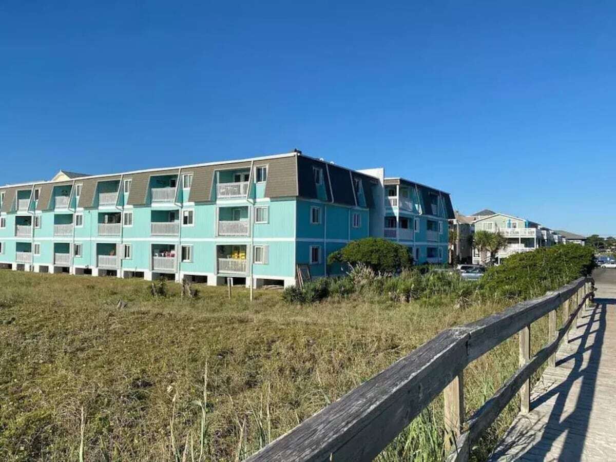 A row of light blue multi-story buildings with balconies, a wooden boardwalk in the foreground, and a clear blue sky.
