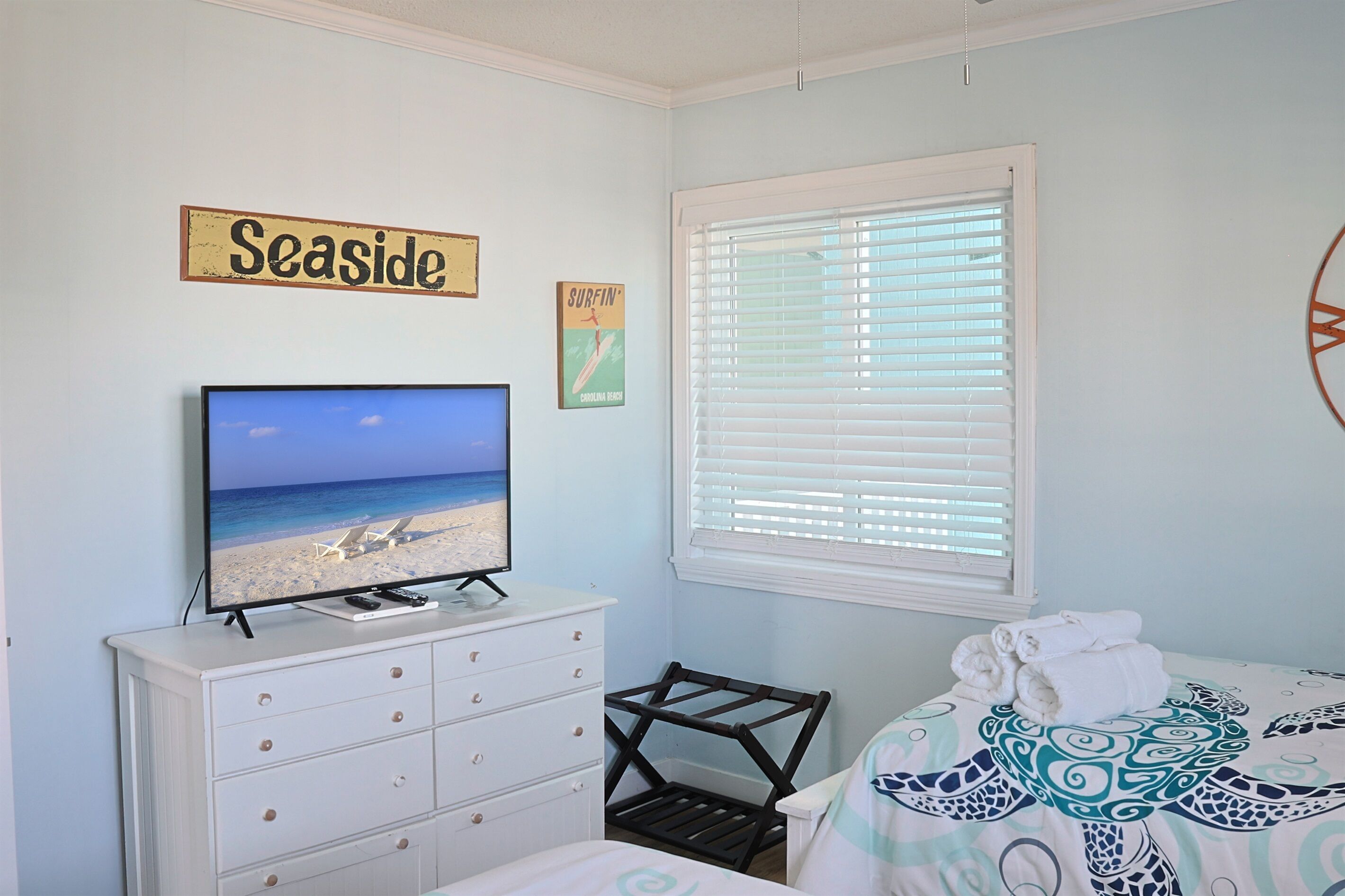 Une chambre avec un téléviseur diffusant une scène de plage, un lit recouvert d’une couvre-lit à motifs, une fenêtre avec des stores, une commode et un panneau indiquant « Au bord de la mer ».