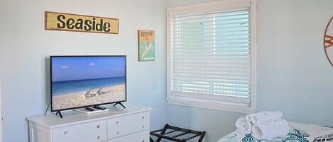 Une chambre avec un téléviseur diffusant une scène de plage, un lit recouvert d’une couvre-lit à motifs, une fenêtre avec des stores, une commode et un panneau indiquant « Au bord de la mer ».