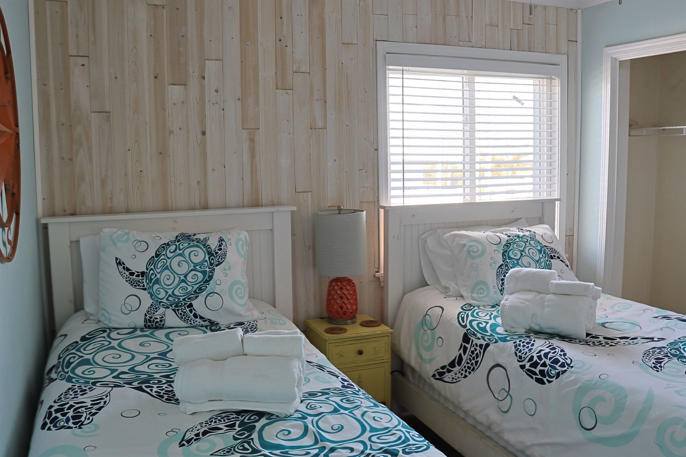 Un dormitorio con dos camas, cada una con ropa de cama y almohadas estampadas, una mesita de noche pequeña con una lámpara y una ventana con persianas.