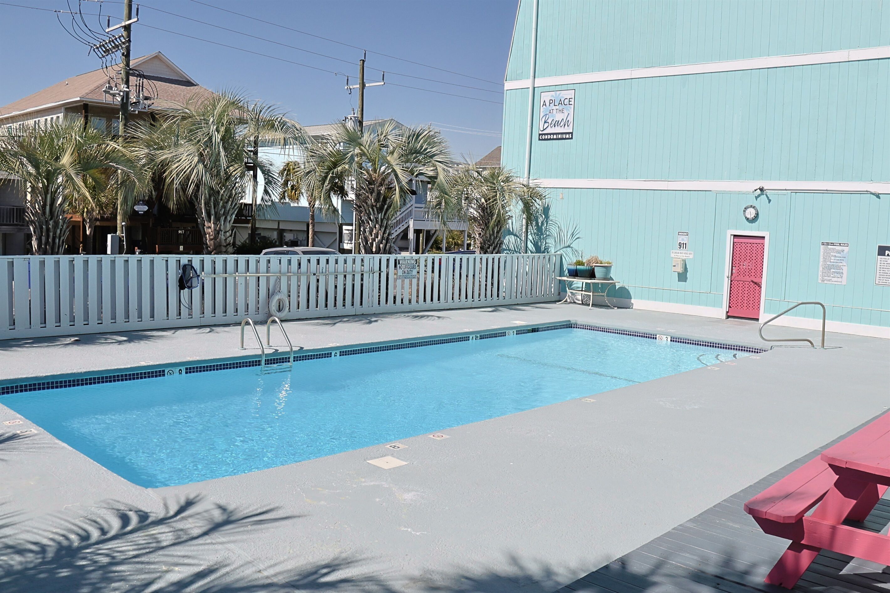 Une piscine entourée d’une clôture blanche, avec une porte rouge et un panneau indiquant : « UN PETIT COIN DE PARADIS SUR LA PLAGE ».