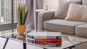 Ein modernes Wohnzimmer mit einem Glastisch, einem grauen Sofa und Blick auf eine Stadtlandschaft durch ein großes Fenster mit Vorhang.