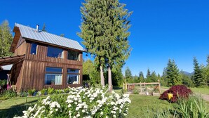 A modern wooden house with large windows and a metal roof, surrounded by lush greenery and a clear blue sky. - Your Lake & Mountain vacation awaits... (Sagle)