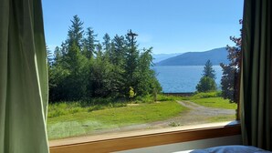 A bedroom with a large window overlooking a lake, surrounded by greenery and a path. - Your Lake & Mountain vacation awaits... (Sagle)