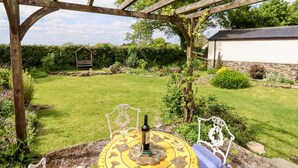 Un jardín con una pérgola, una mesa con mantel amarillo y una botella de vino.
