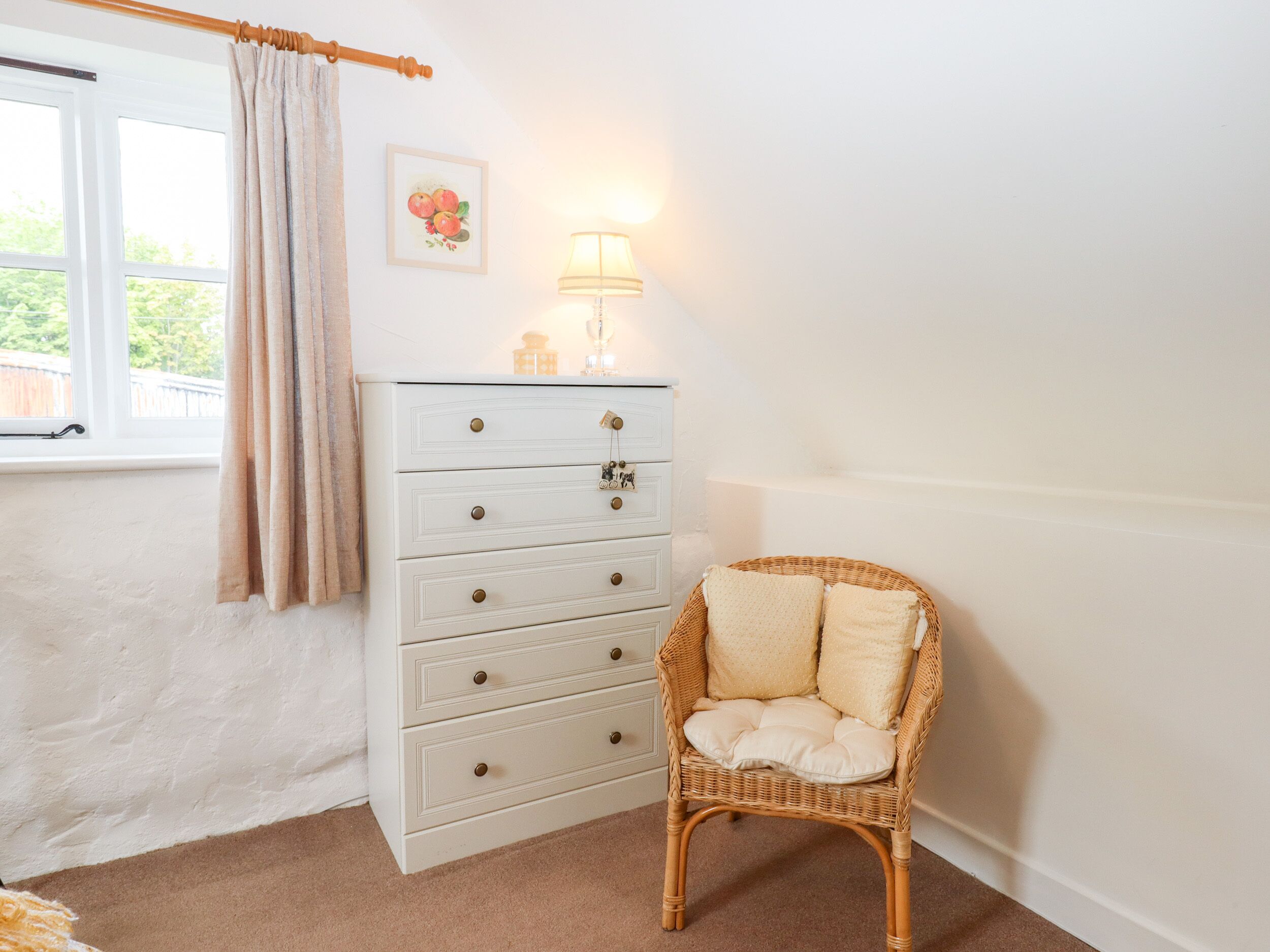 A room with a white dresser, a wicker chair with cushions, a window with curtains, and a wall-mounted lamp.