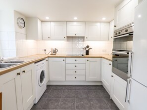 A modern kitchen with white cabinets, a built-in washing machine, a coffee maker, and an oven. - Nuthatch Cottage (Okehampton)