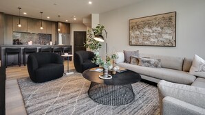 Een moderne woonkamer met een zwarte salontafel, een grijze bank en zwarte fauteuils. Op de achtergrond is een keuken met houten kastjes en een eethoek met een bargedeelte zichtbaar.