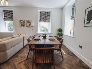 Uma sala de jantar com uma mesa de madeira posta para quatro pessoas, rodeada por cadeiras de madeira. Há duas janelas com cortinas estampadas, um vaso de planta e quadros nas paredes.