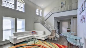 Un salon spacieux avec un canapé d’angle blanc, un tapis coloré, une table basse en verre et un escalier menant à l’étage supérieur.