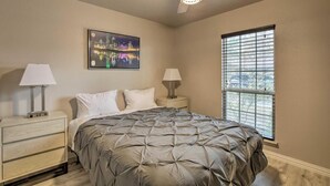 A bedroom with a bed, two bedside tables with lamps, a ceiling fan, a window with blinds, and a cityscape painting on the wall. - Collins Family-Friendly Texas Home with Private Pool! (Garland)