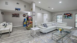A spacious living room with a white sectional sofa, a glass coffee table, and a white couch. There are arcade games, a painting, and a door in the room. - Collins Family-Friendly Texas Home with Private Pool! (Garland)