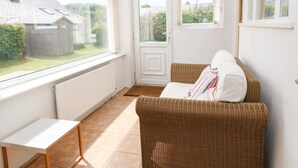 Een lichte kamer met een rieten bank, een klein tafeltje en uitzicht op een huis door grote ramen.