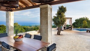 An outdoor patio with a wooden table, wicker chairs, and a fruit bowl. There is a stone pillar, a tree, and a view of the ocean.