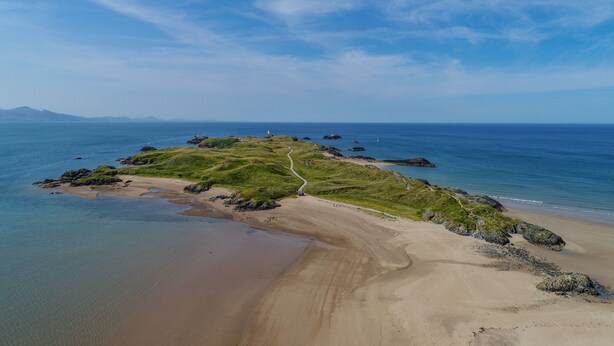 A coastal landscape with a sandy beach, grassy hill, and a winding path. - A Mor (Llanfairpwllgwyngyll)