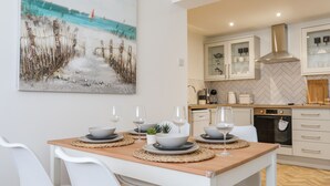 A dining area with a beach-themed painting, a wooden table, white chairs, and a kitchen in the background. - A Mor (Llanfairpwllgwyngyll)