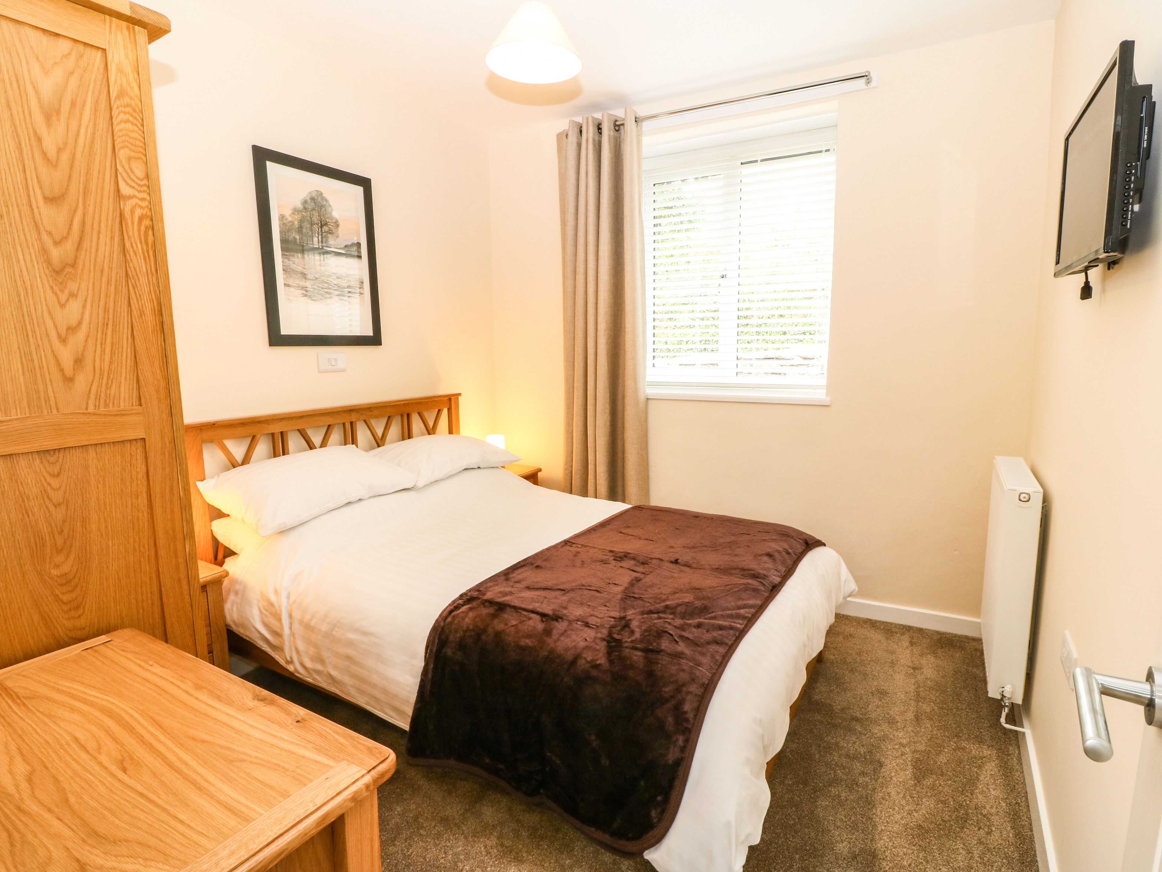 A bedroom with a bed, a wooden desk, a framed picture on the wall, a window with curtains, and a radiator.