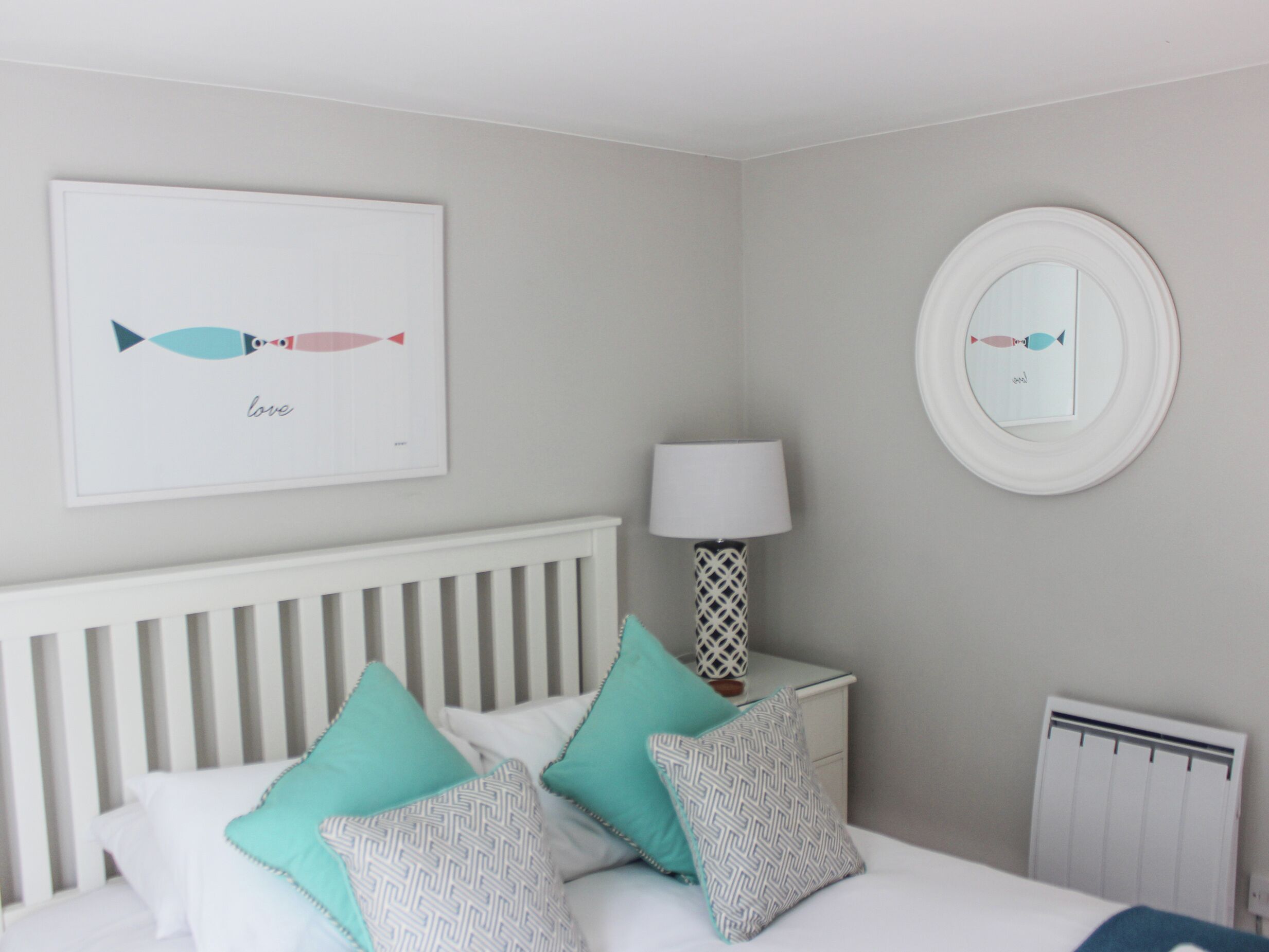 Un dormitorio con cama, almohadas, una mesita de noche con lámpara, un espejo en la pared y un cuadro enmarcado.