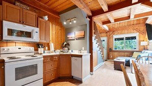 A spacious kitchen with wooden cabinets, a microwave, stove, and a dishwasher. There is a dining area with a table and chairs, and a window providing a view of the outdoors. - Bear Coast | Cabin w/ Hot Tub, Patio, & Waterview (Ucluelet)