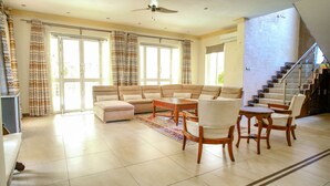 Amplia sala de estar con sofá y sillones a juego en tono beige, y una mesa de centro de madera. Un ventanal grande permite que la luz natural ilumine la habitación, y se observa un ventilador de techo.