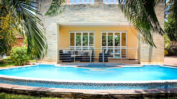 Una piscina con borde de azulejos, una zona de estar cubierta con sofá y sillas, y un muro de piedra al fondo.