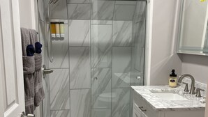 Ein Badezimmer mit einer gläsernen Duschkabine, einer weißen Toilette, einem Waschbecken mit Marmorplatte und einem dunklen Teppich auf dem Boden.