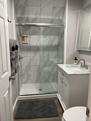 Un baño con ducha envuelta en vidrio, inodoro blanco, lavamanos con encimera de mármol y alfombra oscura en el suelo.