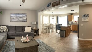 Ein Wohnzimmer mit grauem Ecksofa, einem Couchtisch mit Dekoration, einem Bild an der Wand und einem Essbereich mit Holztisch und Stühlen.