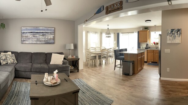 A living room with a gray sectional sofa, a coffee table with decorative items, a painting on the wall, and a dining area with a wooden table and chairs. - Spacious Home for 14 Guests Near Lake Winnisquam – Close to Everything! (Sanbornton)