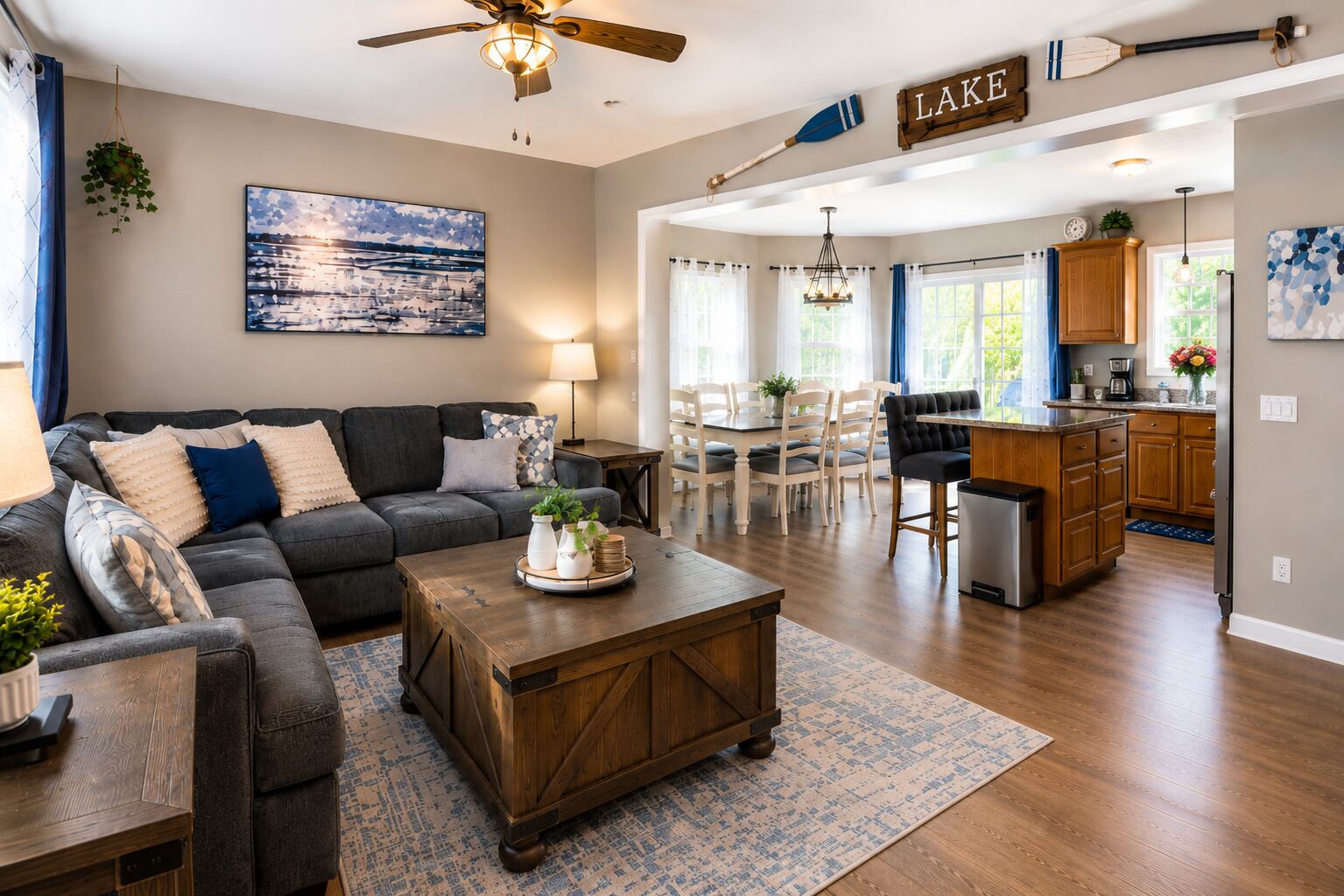 A spacious living room with a dark grey sofa, a wooden coffee table, and a dining area with white chairs. A kitchen with wooden cabinets and a bar area is visible in the background.
