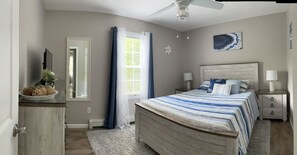 Une chambre à coucher comprenant un lit, des tables de chevet, un ventilateur de plafond, une fenêtre avec des rideaux et une œuvre d’art accrochée au mur.
